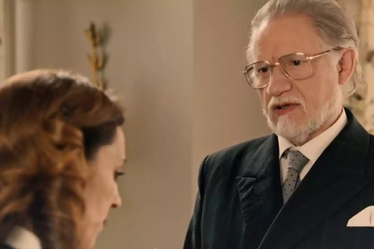Damián amb barba i ulleres parla seriosament amb Manuela de cabells castanys en un ambient interior elegant. Captura de 'somnis de llibertat' Damián amb barba i ulleres parla seriosament amb Manuela de cabells castanys en un ambient interior elegant. Captura de 'somnis de llibertat'