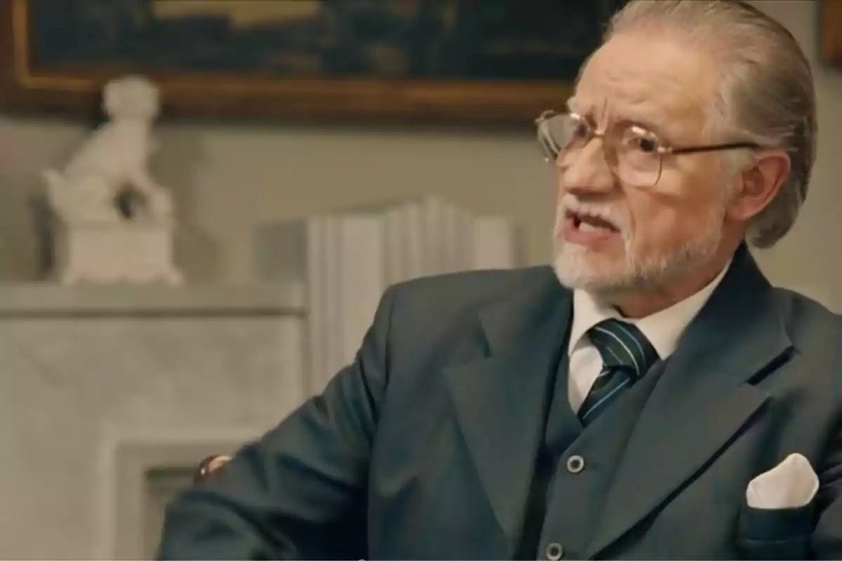 Damián con gafas y barba canosa vestido con traje oscuro y corbata a rayas sentado en una habitación elegante. Captura de 'sueños de libertad' Damián con gafas y barba canosa vestido con traje oscuro y corbata a rayas sentado en una habitación elegante. Captura de 'sueños de libertad'