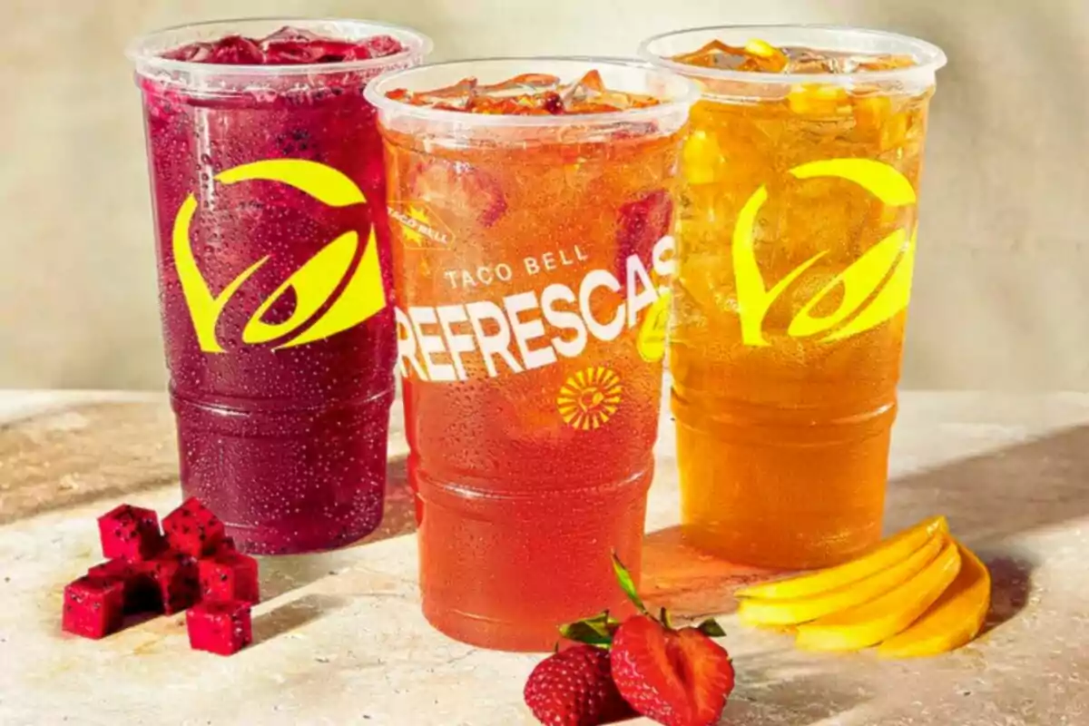 Tres vasos grandes de bebidas refrescantes de colores rojo, naranja y amarillo con hielo, acompañados de trozos de fruta como fresa, mango y pitaya, sobre una superficie clara Tres vasos grandes de bebidas refrescantes de colores rojo, naranja y amarillo con hielo, acompañados de trozos de fruta como fresa, mango y pitaya, sobre una superficie clara