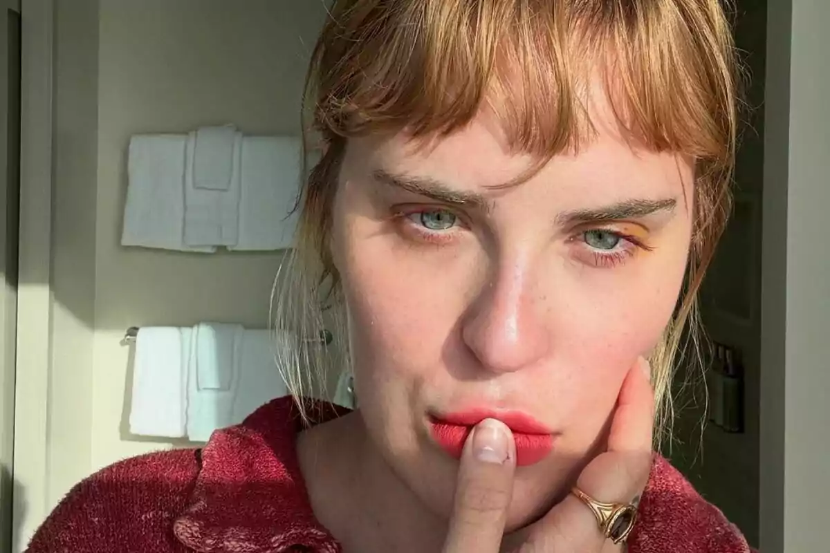 Person with blonde hair and light eyes touching their lips with a finger in a bathroom lit by natural light, with white towels hanging on the wall in the background. Person with blonde hair and light eyes touching their lips with a finger in a bathroom lit by natural light, with white towels hanging on the wall in the background.