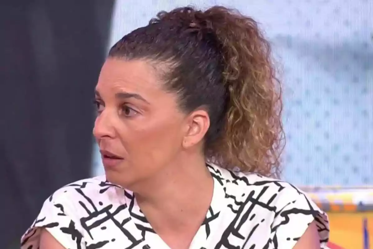 Plano corto de Tamara, hermana de Michu, con el cabello rizado y recogido en una coleta mirando hacia la izquierda con expresión seria, vestida con blusa blanca de líneas negras en un set de televisión. Plano corto de Tamara, hermana de Michu, con el cabello rizado y recogido en una coleta mirando hacia la izquierda con expresión seria, vestida con blusa blanca de líneas negras en un set de televisión.