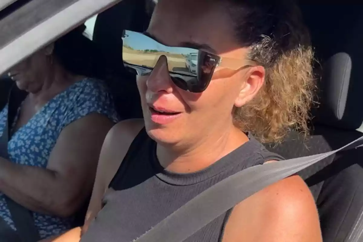 Tamara amb ulleres de sol grans i samarreta de tirants negra asseguda al seient del copilot d’un cotxe amb el cinturó de seguretat posat i una altra persona al volant Tamara amb ulleres de sol grans i samarreta de tirants negra asseguda al seient del copilot d’un cotxe amb el cinturó de seguretat posat i una altra persona al volant