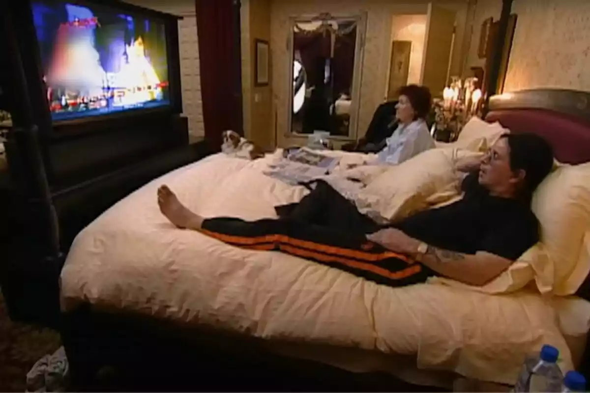 Ozzy Osbourne con más personas de su familia y un perro están acostados en una cama grande viendo la televisión en una habitación decorada con tonos cálidos y un espejo al fondo Ozzy Osbourne con más personas de su familia y un perro están acostados en una cama grande viendo la televisión en una habitación decorada con tonos cálidos y un espejo al fondo