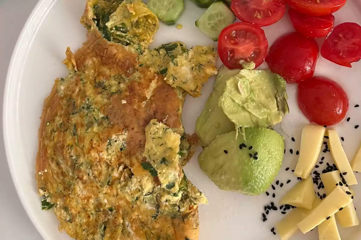 Omelette with herbs served with slices of cucumber, cherry tomatoes, avocado, and strips of cheese with black seeds on a white plate. Omelette with herbs served with slices of cucumber, cherry tomatoes, avocado, and strips of cheese with black seeds on a white plate.