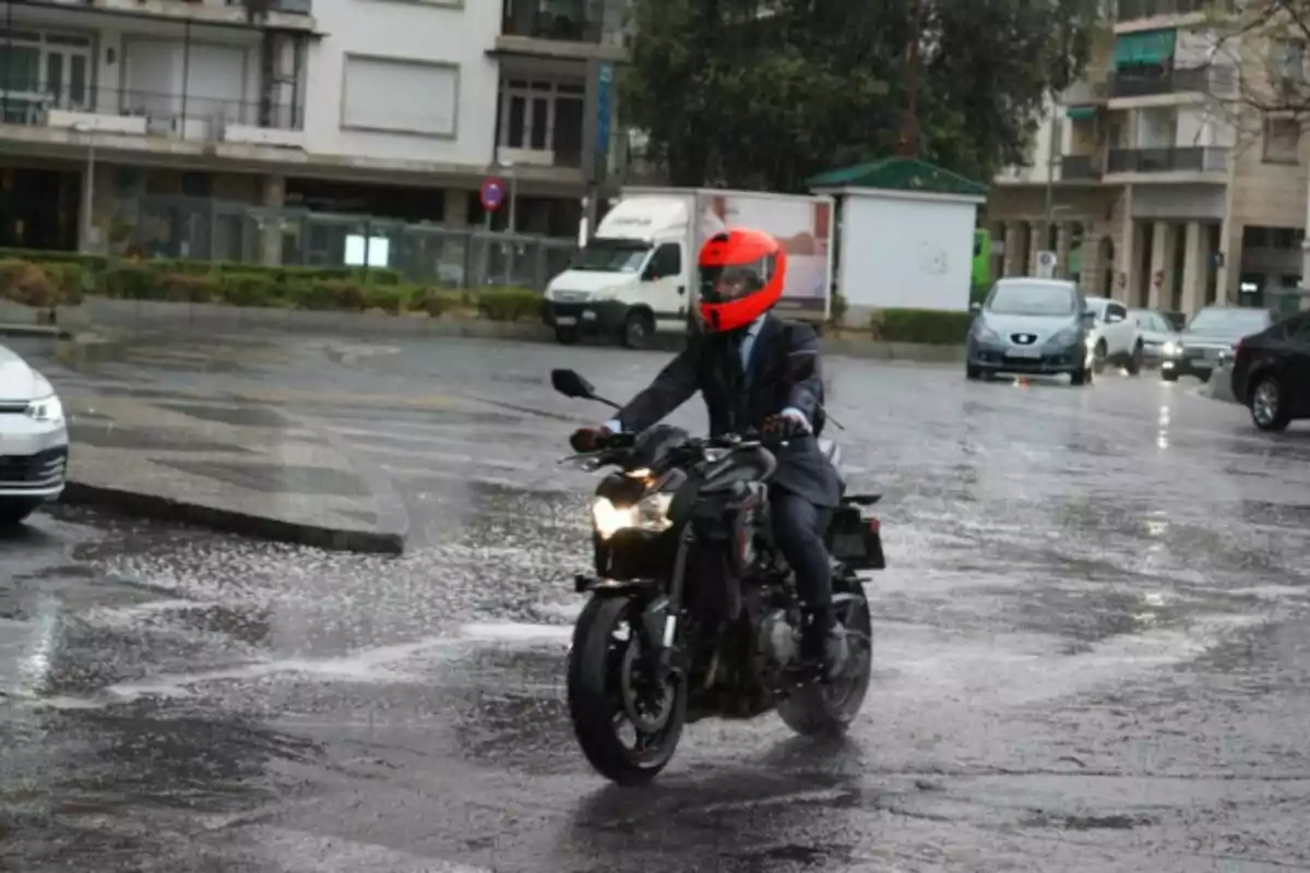 Persona amb casc vermell conduint una motocicleta per un carrer mullat per la pluja envoltat de cotxes i edificis. Persona amb casc vermell conduint una motocicleta per un carrer mullat per la pluja envoltat de cotxes i edificis.