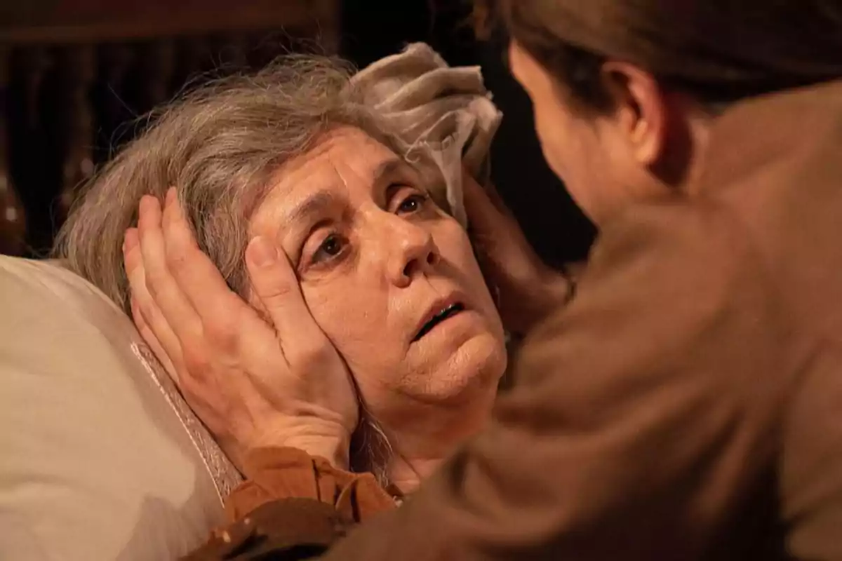 Raimunda estirada en un llit és sostinguda pel rostre amb totes dues mans per una altra persona que la mira de prop mostrant una expressió de preocupació i tendresa. Captura de 'Valle Salvaje' Raimunda estirada en un llit és sostinguda pel rostre amb totes dues mans per una altra persona que la mira de prop mostrant una expressió de preocupació i tendresa. Captura de 'Valle Salvaje'