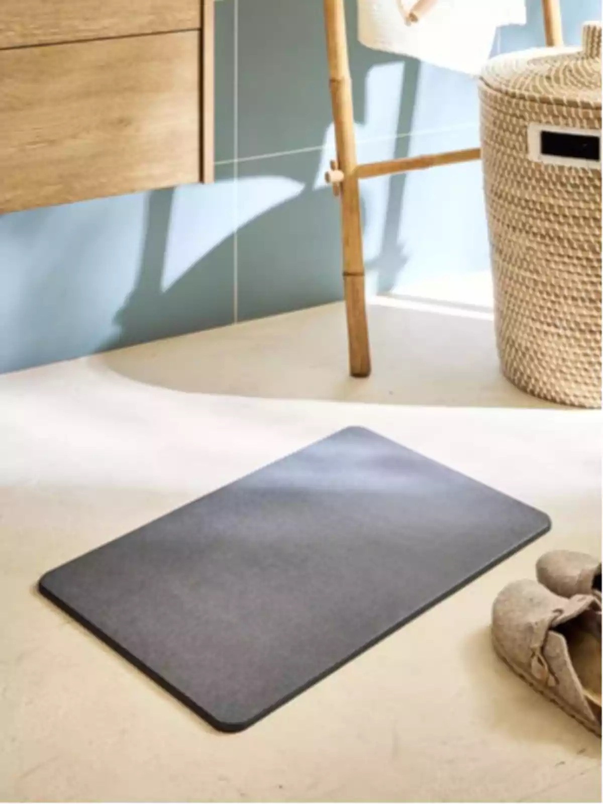 Estora rectangular grisa al terra d’un bany al costat d’un cistell de vímet i unes sabatilles beix.