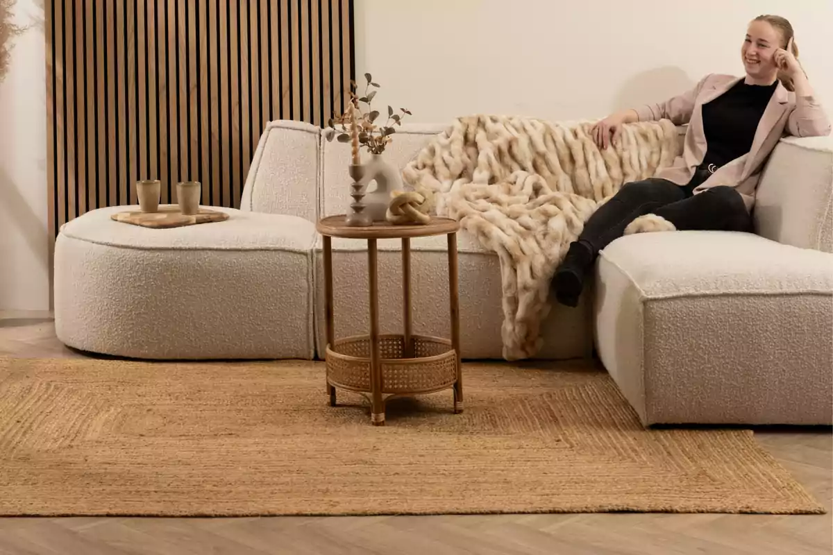 Mujer sentada en un sofá blanco con manta de peluche, mesa auxiliar de madera y decoración minimalista en una sala acogedora Mujer sentada en un sofá blanco con manta de peluche, mesa auxiliar de madera y decoración minimalista en una sala acogedora