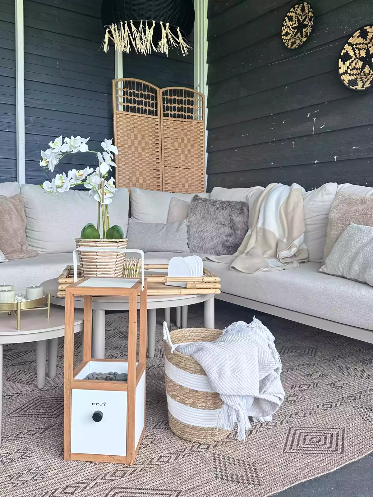 Sala de estar acogedora con sofá en tonos claros, cojines, mantas, una mesa de centro con bandeja y velas, una lámpara colgante negra, una pantalla de mimbre y decoración de pared tejida Sala de estar acogedora con sofá en tonos claros, cojines, mantas, una mesa de centro con bandeja y velas, una lámpara colgante negra, una pantalla de mimbre y decoración de pared tejida