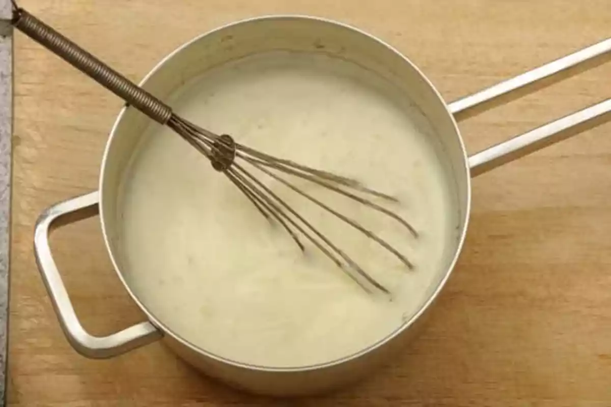 Cazo con bechamel y batidor de varillas sobre una tabla de madera Cazo con bechamel y batidor de varillas sobre una tabla de madera