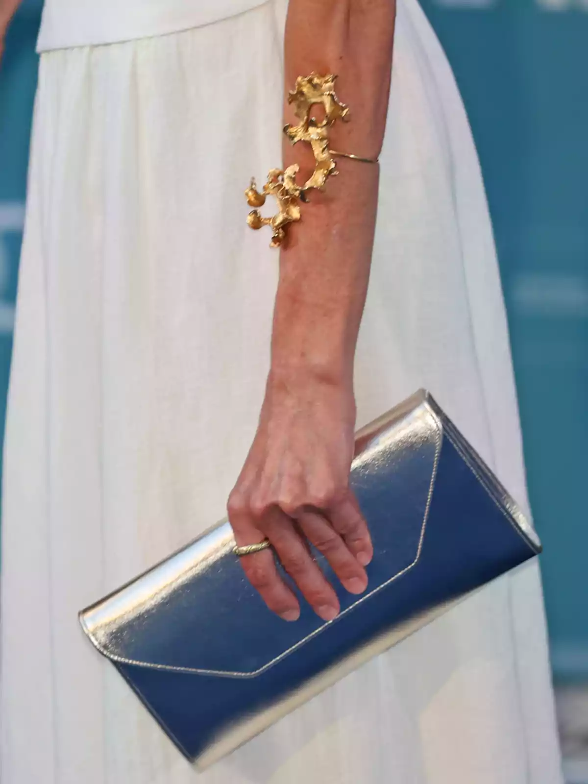 La reina Letícia amb vestit blanc sostenint una bossa metàl·lica platejada i portant una braçalet daurat cridaner al braç La reina Letícia amb vestit blanc sostenint una bossa metàl·lica platejada i portant una braçalet daurat cridaner al braç