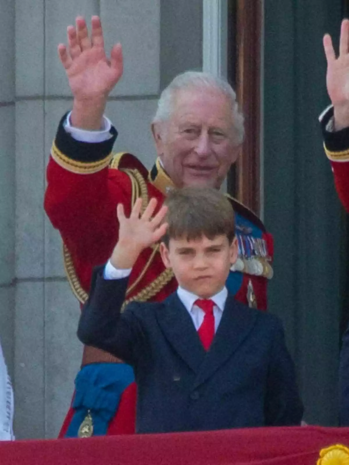 Carles III amb uniforme vermell i el príncep Louis amb vestit fosc i corbata vermella saluden des d’un balcó Carles III amb uniforme vermell i el príncep Louis amb vestit fosc i corbata vermella saluden des d’un balcó