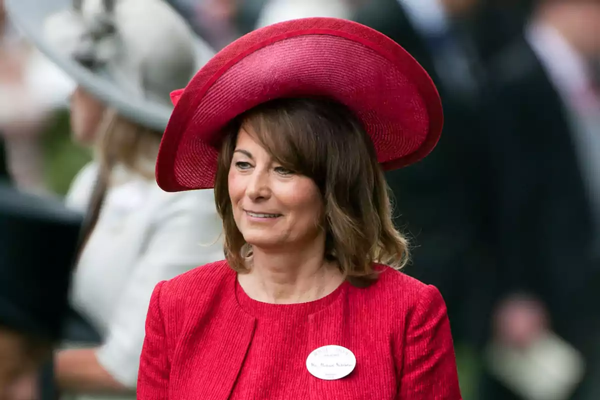 Carole Middleton con sombrero y vestido rojo sonríe mientras asiste a un evento formal al aire libre Carole Middleton con sombrero y vestido rojo sonríe mientras asiste a un evento formal al aire libre