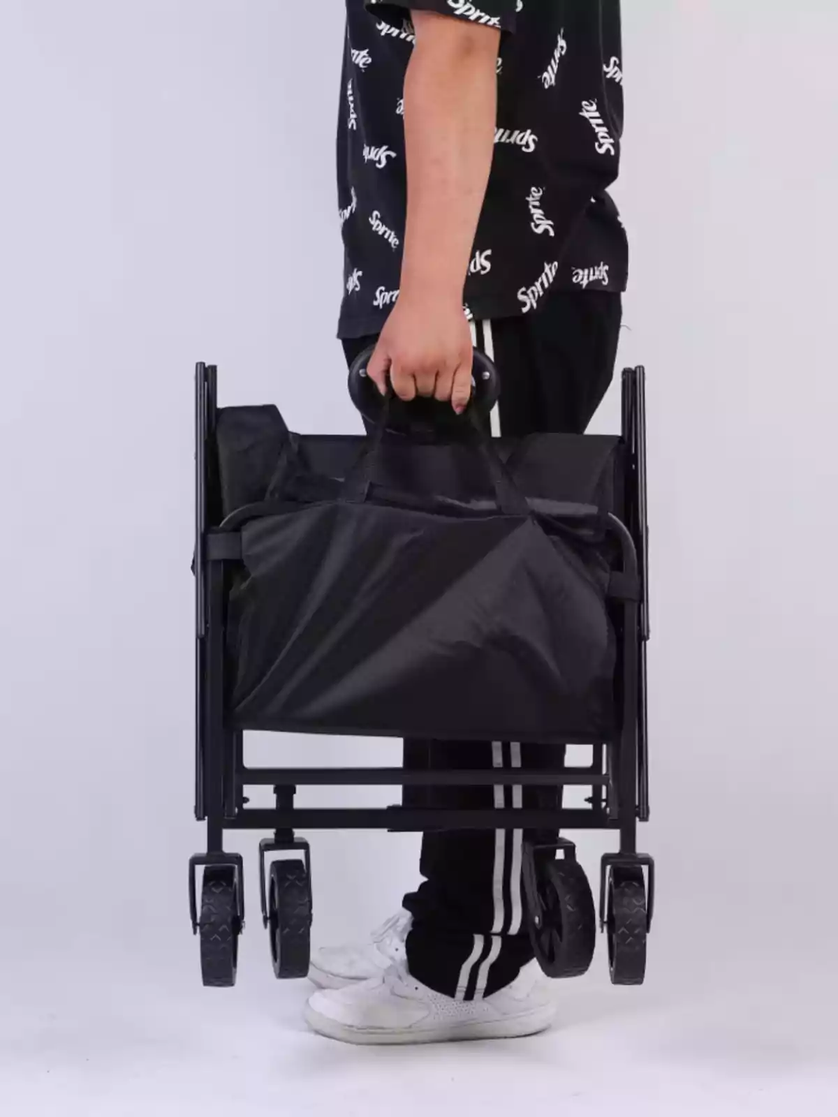 Persona sostenint un carro plegable negre amb rodes, vestint pantalons esportius i vambes blanques Persona sostenint un carro plegable negre amb rodes, vestint pantalons esportius i vambes blanques