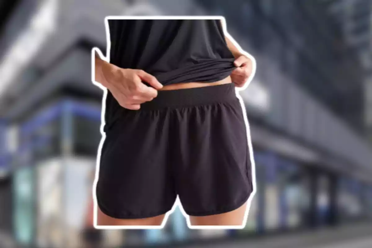 Persona amb pantalons curts esportius negres aixecant lleugerament la samarreta davant d’un fons urbà desenfocat Persona amb pantalons curts esportius negres aixecant lleugerament la samarreta davant d’un fons urbà desenfocat