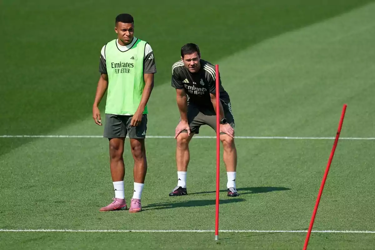 Dues persones amb roba esportiva del Reial Madrid són en un camp de futbol durant un entrenament al costat d’uns pals vermells.