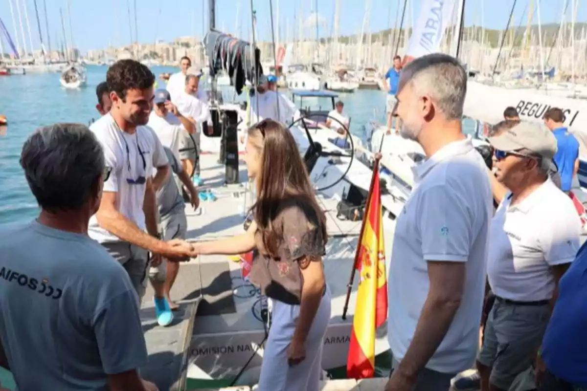 Una dona saluda un home en un vaixell envoltats de diverses persones en un port amb banderes i velers al fons. Una dona saluda un home en un vaixell envoltats de diverses persones en un port amb banderes i velers al fons.