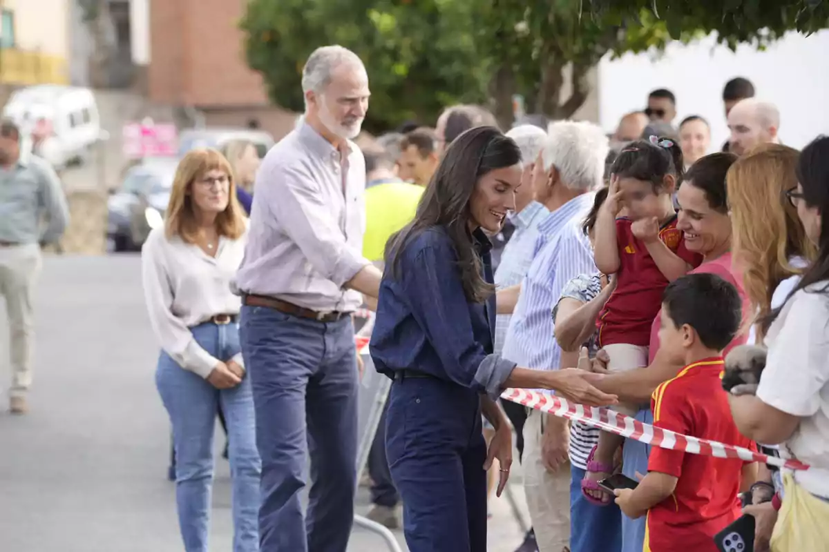 La reina Letícia somrient saluda un grup de persones darrere d’una cinta de seguretat en un esdeveniment a l’aire lliure, mentre Felip VI i més assistents observen l’escena.