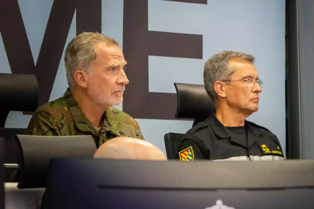 Felipe VI and another man in a military and civil protection uniform sitting in a control room, alertly watching something off-camera. Felipe VI and another man in a military and civil protection uniform sitting in a control room, alertly watching something off-camera.