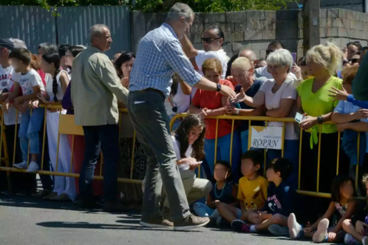 A man greets a group of people who are behind a yellow fence while several children sit on the ground in front of him. A man greets a group of people who are behind a yellow fence while several children sit on the ground in front of him.