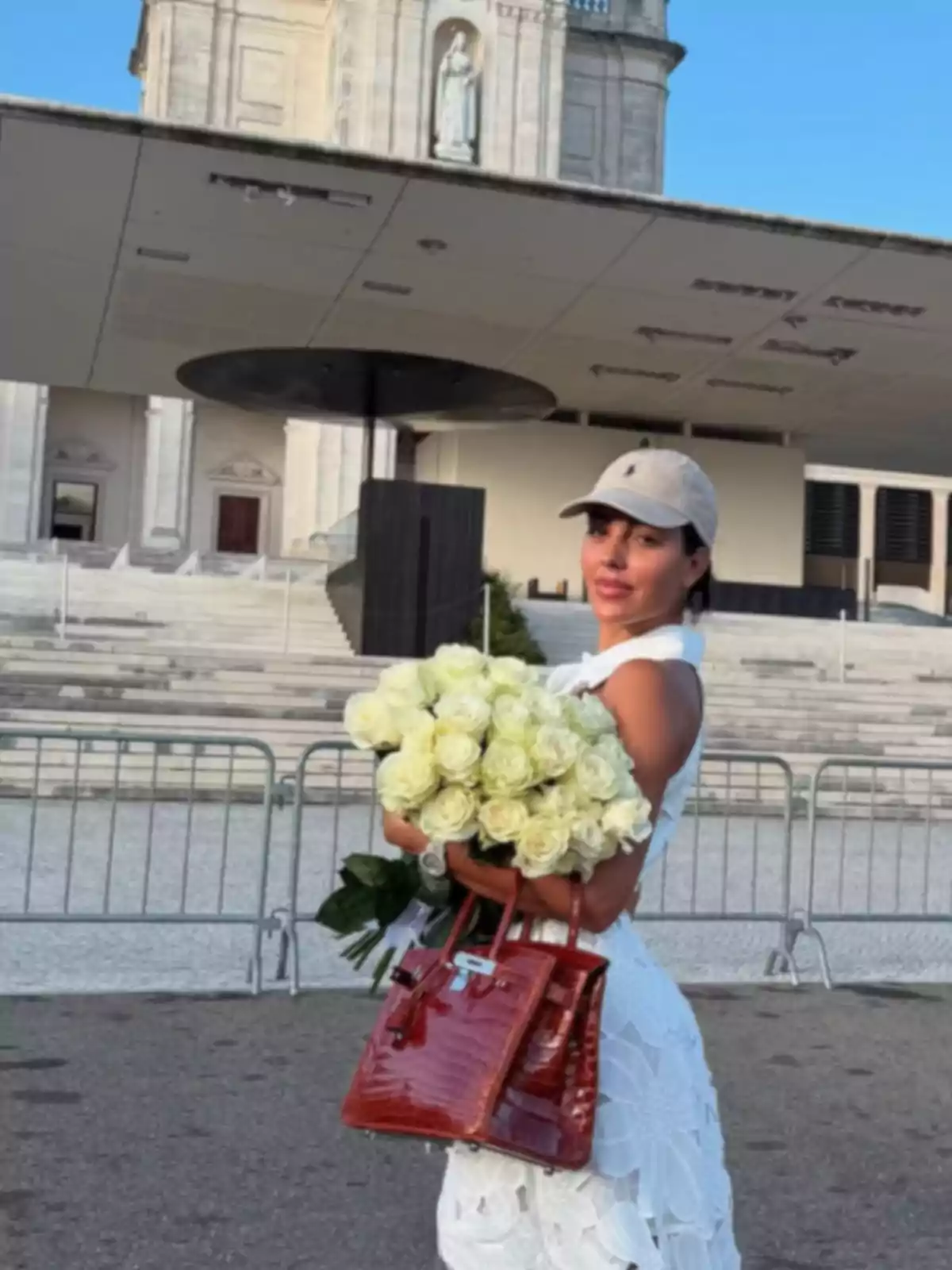 Dona vestida de blanc amb gorra clara sostenint un ram de roses blanques i una bossa vermella davant d’un edifici religiós Dona vestida de blanc amb gorra clara sostenint un ram de roses blanques i una bossa vermella davant d’un edifici religiós