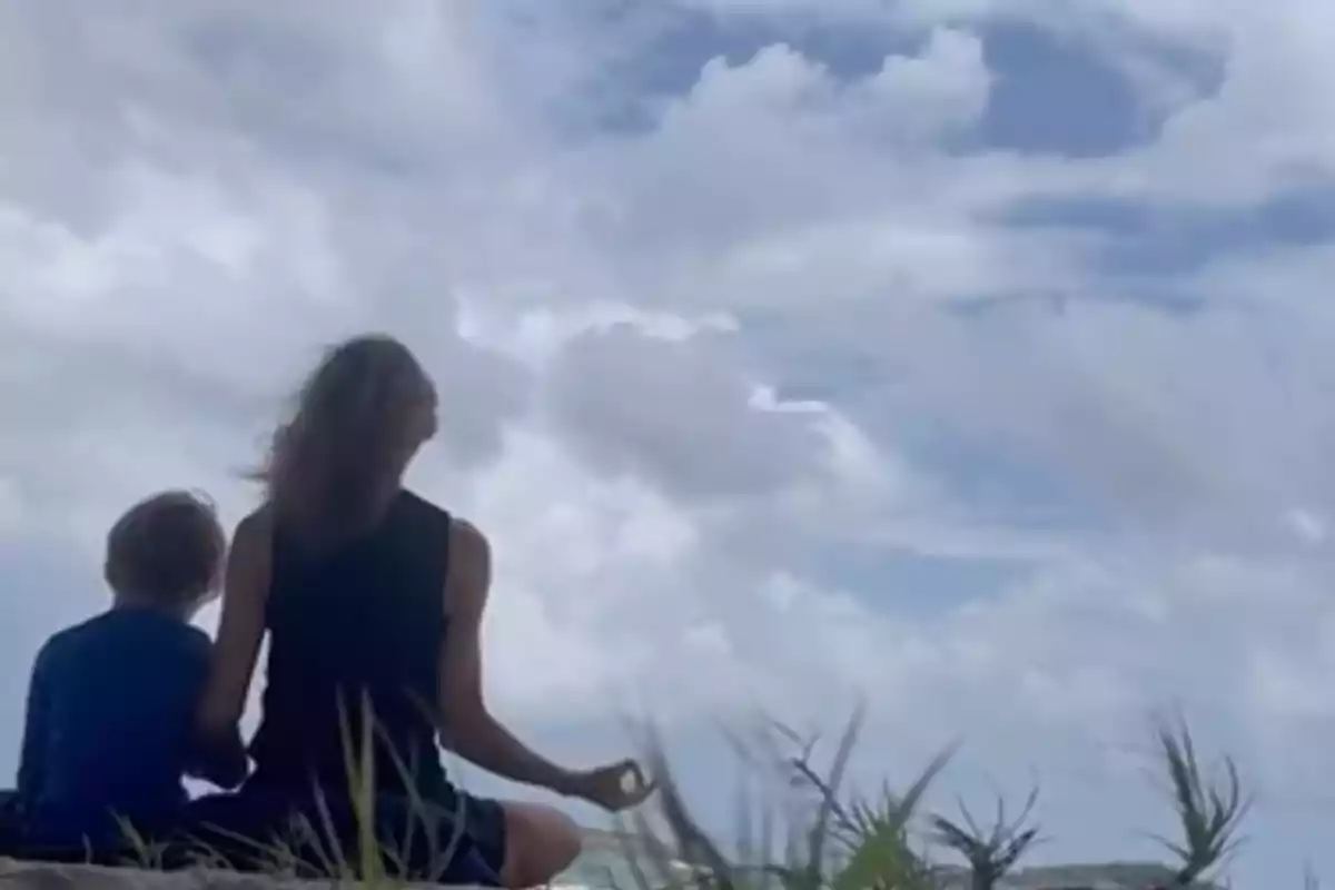 Alejandra Silva y su hijo sentados de espaldas en el suelo mirando el cielo nublado rodeadas de vegetación baja