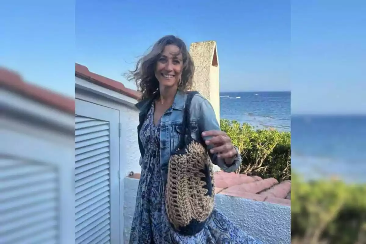 Mujer sonriente con vestido azul y chaqueta de mezclilla, sosteniendo un bolso tejido, de pie al aire libre junto al mar y una casa blanca