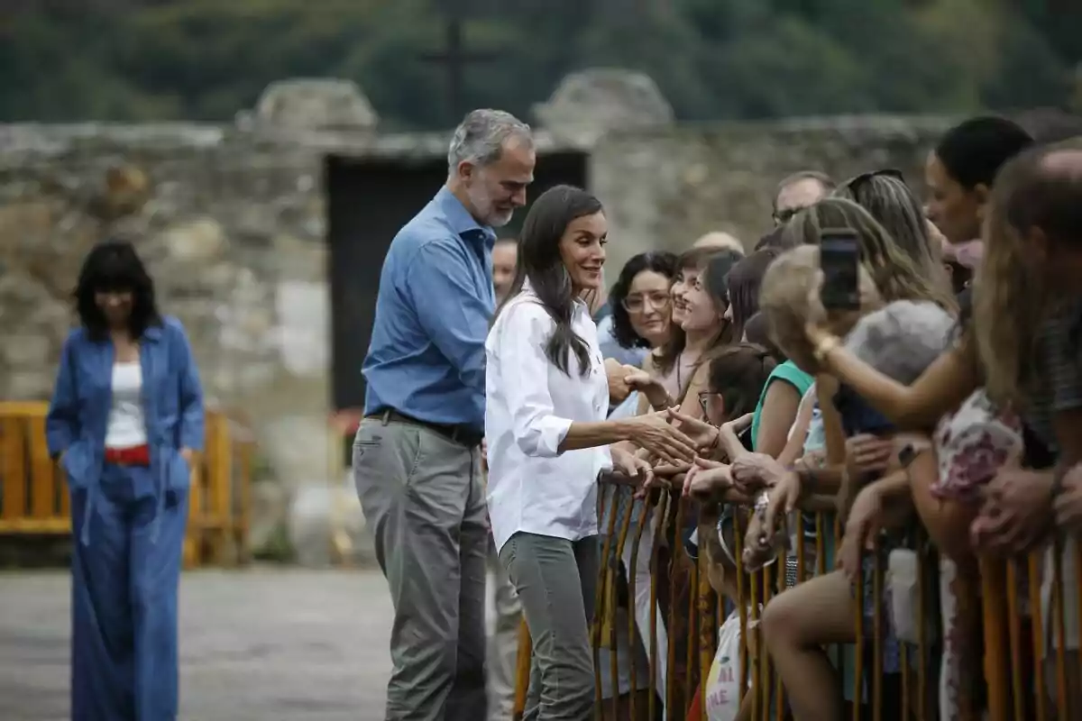 Una parella saluda i dona la mà a diverses persones que són darrere d’una tanca en un entorn a l’aire lliure amb una paret de pedra al fons. Una parella saluda i dona la mà a diverses persones que són darrere d’una tanca en un entorn a l’aire lliure amb una paret de pedra al fons.