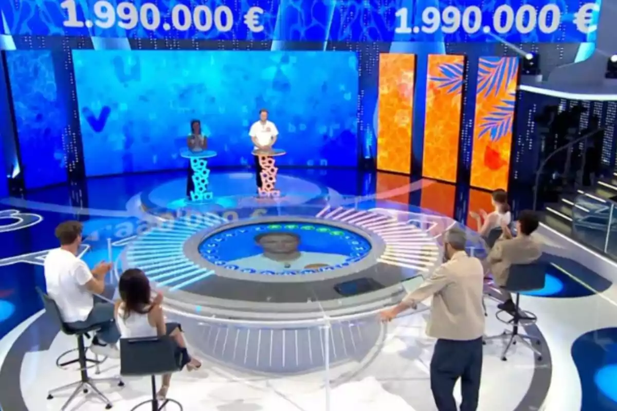 Un plató de concurs televisiu amb dos concursants drets davant d’atrils i diverses persones assegudes al seu voltant mentre aplaudeixen, amb una gran pantalla blava de fons que mostra una quantitat de diners. Un plató de concurs televisiu amb dos concursants drets davant d’atrils i diverses persones assegudes al seu voltant mentre aplaudeixen, amb una gran pantalla blava de fons que mostra una quantitat de diners.