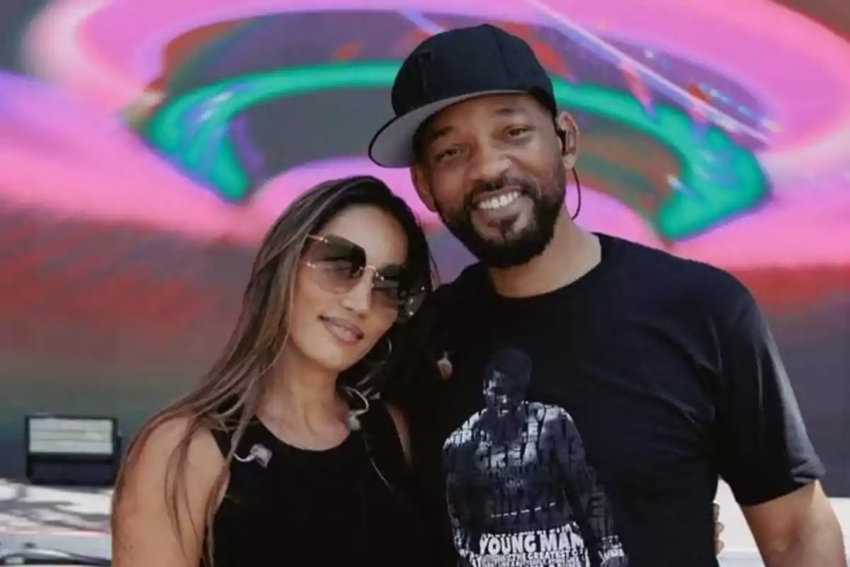 Two people pose smiling in front of a colorful background with neon lights; one of them is wearing sunglasses and the other a black cap. Two people pose smiling in front of a colorful background with neon lights; one of them is wearing sunglasses and the other a black cap.