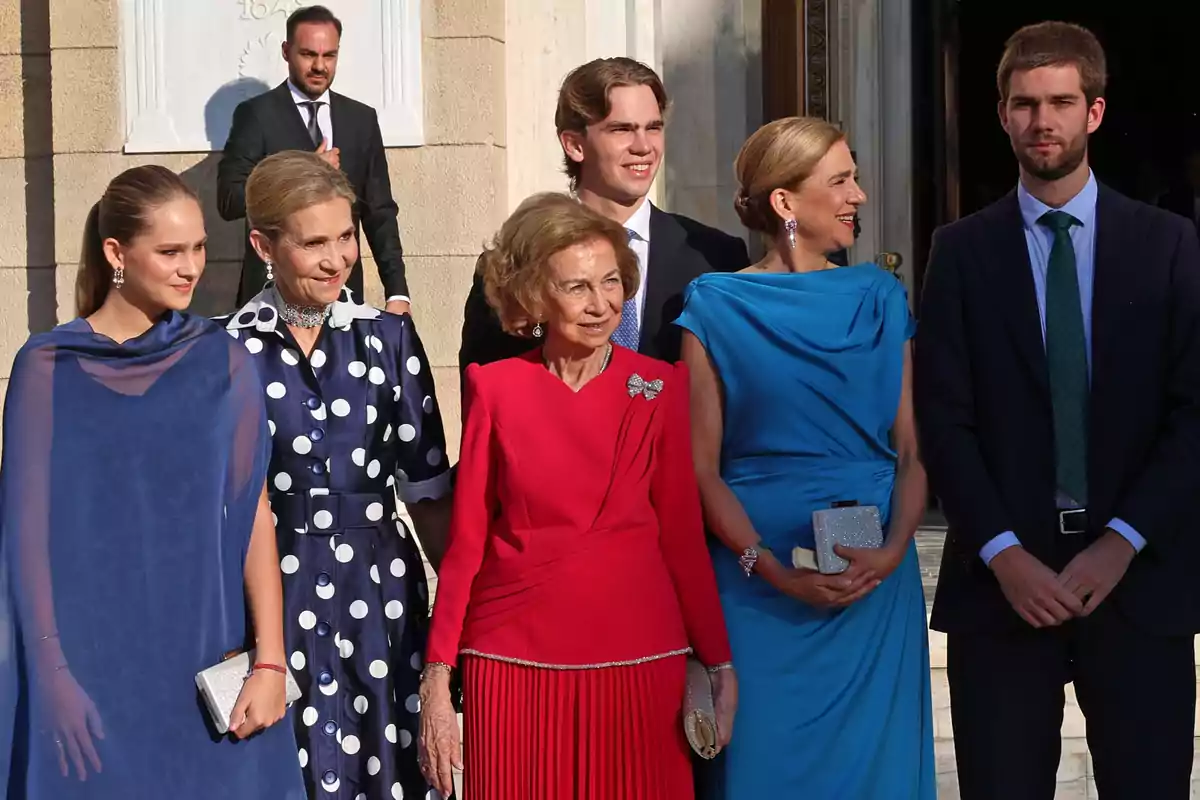 A group of elegantly dressed people posing together outdoors in front of a building, some of them smiling and wearing formal dresses in shades of blue and red. A group of elegantly dressed people posing together outdoors in front of a building, some of them smiling and wearing formal dresses in shades of blue and red.