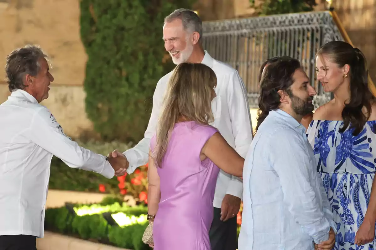 Koldo Royo, wearing a white jersey, shakes hands with a smiling Felipe VI while two couples chat beside them in an outdoor setting with plants and flowers in the background. Koldo Royo, wearing a white jersey, shakes hands with a smiling Felipe VI while two couples chat beside them in an outdoor setting with plants and flowers in the background.