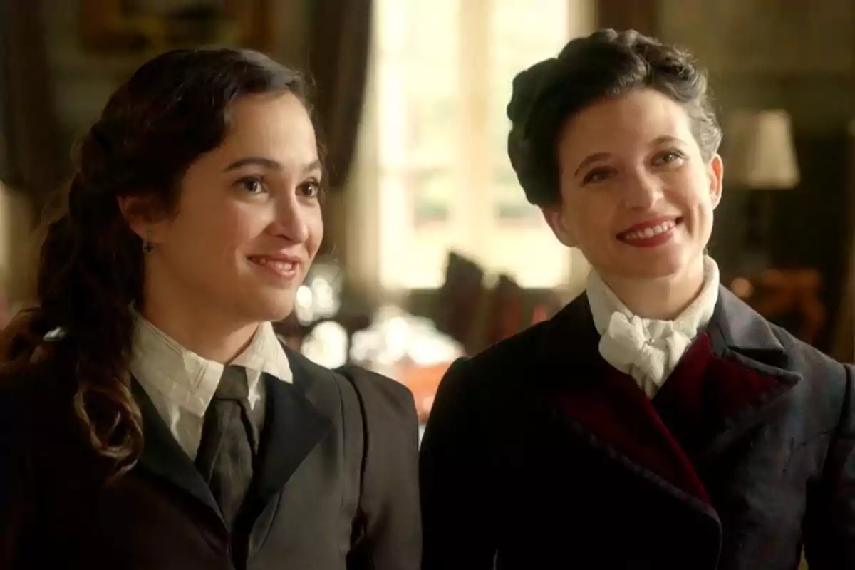 La Catalina i la Martina somriuen vestides amb roba d’època en un ambient elegant i càlid. Captura de ‘La Promesa’