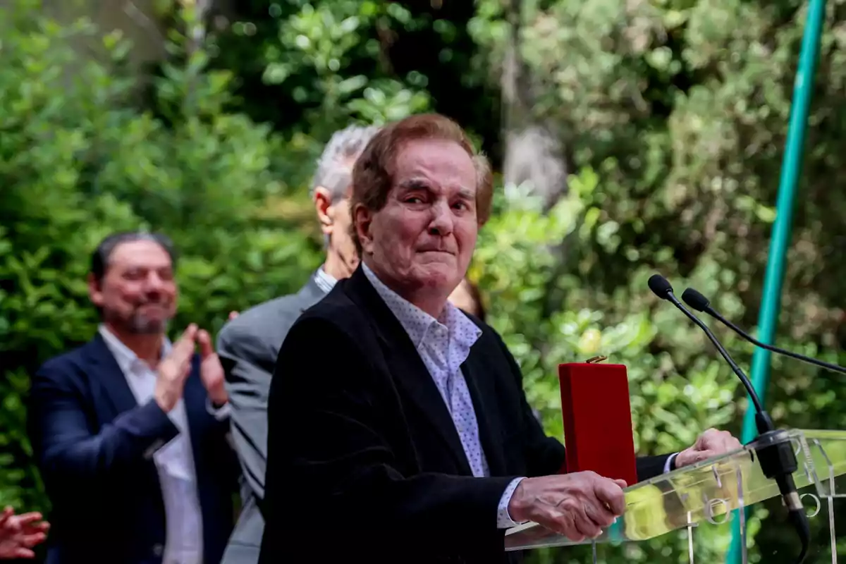 Manuel de la Calva standing in front of a lectern with microphones, holding a red box while several people applaud in the background in an outdoor setting with vegetation. Manuel de la Calva standing in front of a lectern with microphones, holding a red box while several people applaud in the background in an outdoor setting with vegetation.