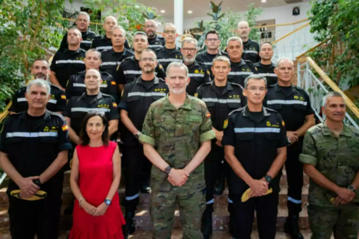 Grupo de personas con uniformes militares y de protección civil posando en una escalera junto a una mujer vestida de rojo en un entorno interior con plantas Grupo de personas con uniformes militares y de protección civil posando en una escalera junto a una mujer vestida de rojo en un entorno interior con plantas