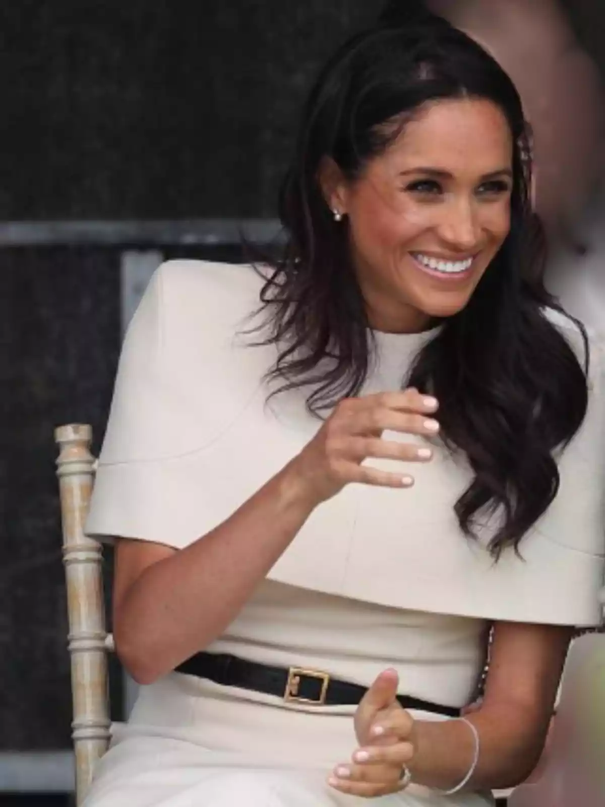 Mujer de cabello oscuro y largo sentada en una silla de madera, sonriente y vestida con un conjunto color crema Mujer de cabello oscuro y largo sentada en una silla de madera, sonriente y vestida con un conjunto color crema