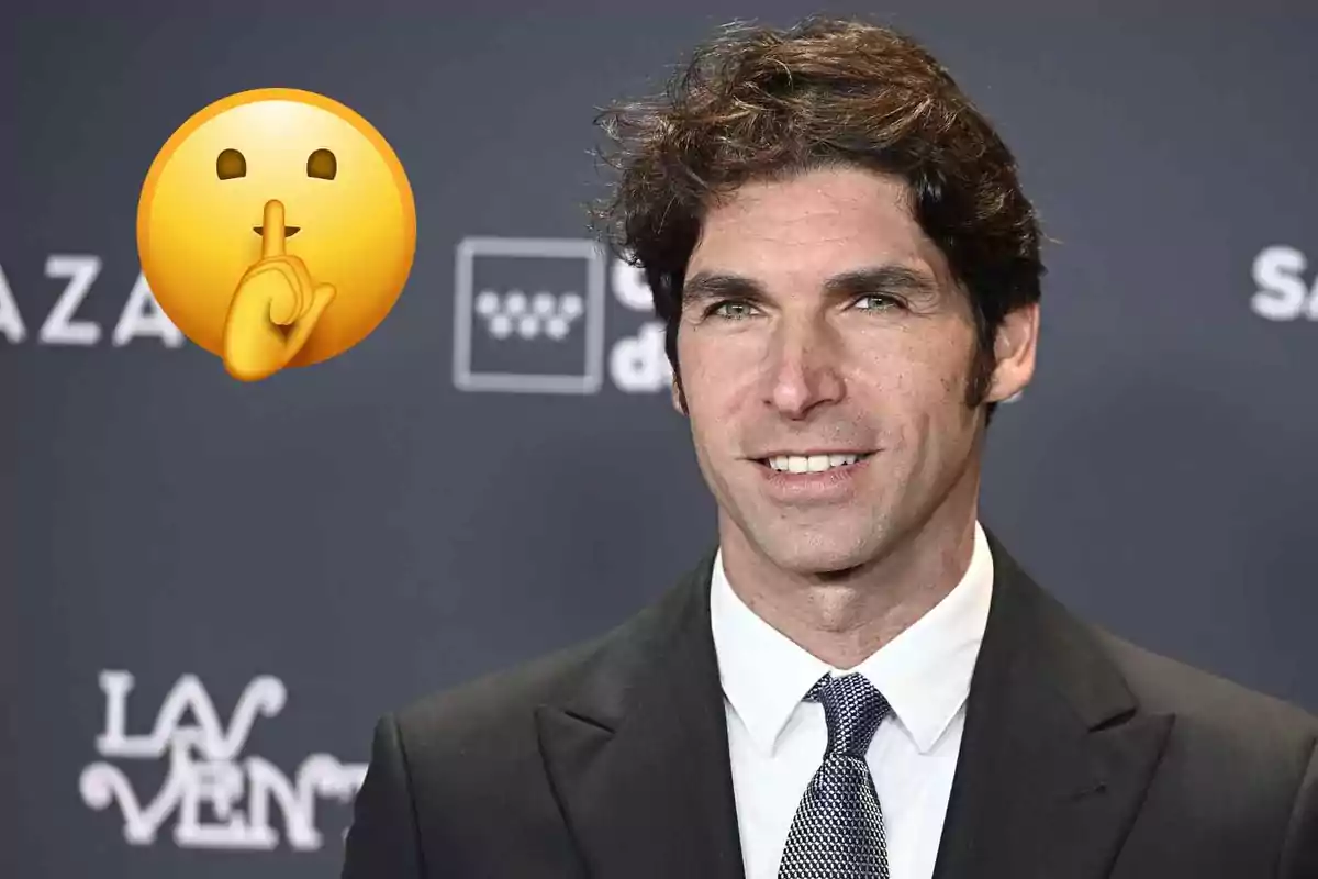 Cayetano Rivera de cabello oscuro y rizado con traje y corbata posando frente a un fondo de evento, acompañado de un emoji de silencio