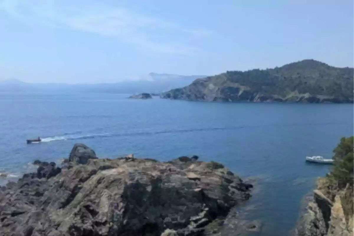 Vista de un paisaje costero con rocas en primer plano, un mar azul tranquilo, una lancha y un barco, y montañas al fondo bajo un cielo despejado Vista de un paisaje costero con rocas en primer plano, un mar azul tranquilo, una lancha y un barco, y montañas al fondo bajo un cielo despejado