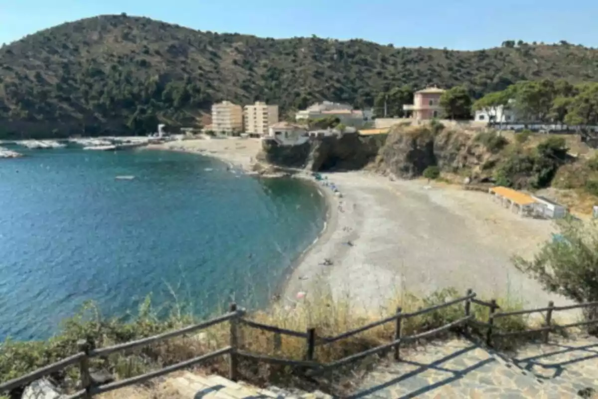 Vista de una playa de aguas tranquilas y arena clara rodeada de colinas y edificios residenciales con una barandilla de madera en primer plano Vista de una playa de aguas tranquilas y arena clara rodeada de colinas y edificios residenciales con una barandilla de madera en primer plano