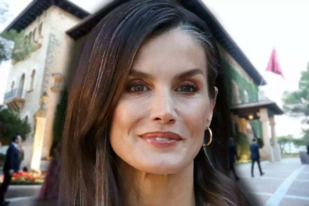 La reina Letizia con arete dorado sonríe levemente frente a un edificio elegante con bandera roja al fondo