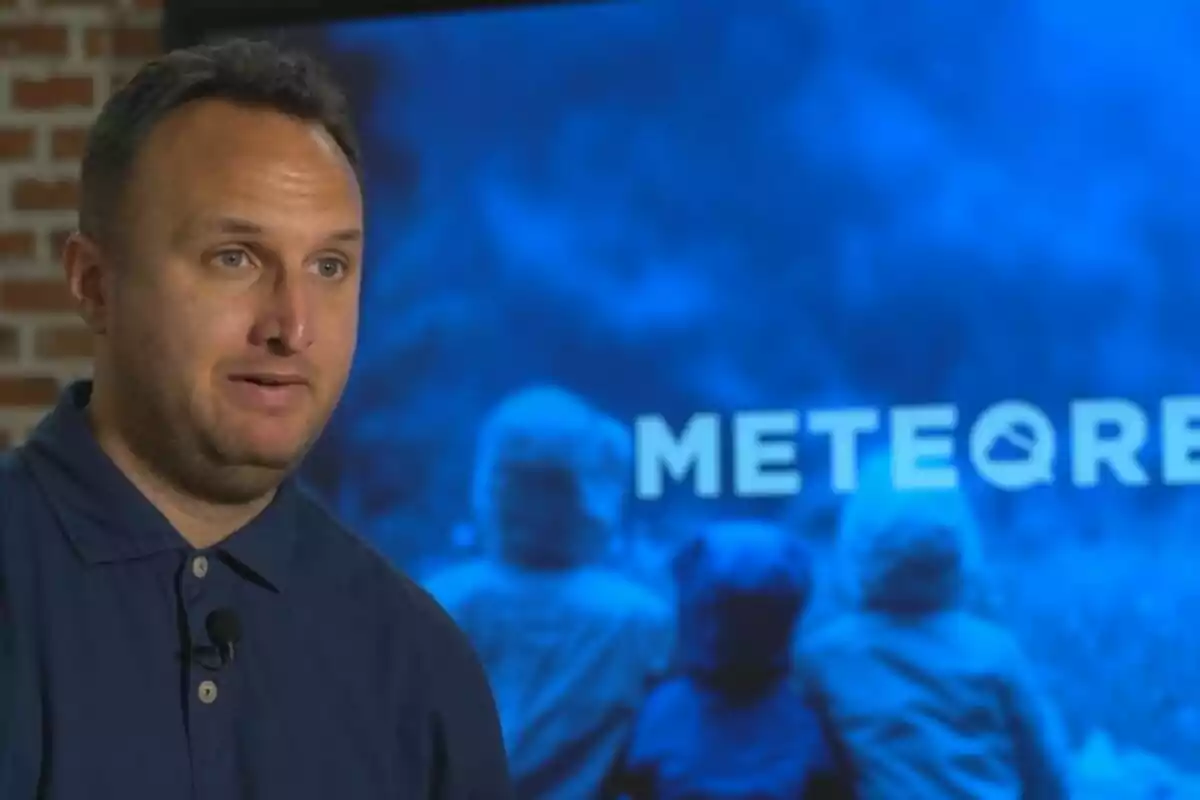 Hombre con camisa azul hablando frente a una pantalla azul que muestra la palabra METEORE y la silueta de varias personas de espaldas. Hombre con camisa azul hablando frente a una pantalla azul que muestra la palabra METEORE y la silueta de varias personas de espaldas.