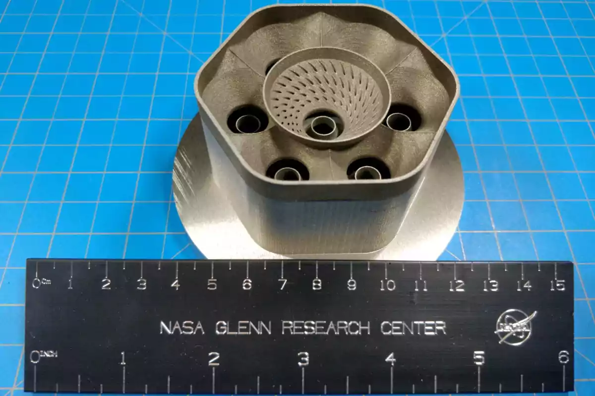 Peça metàl·lica impresa en 3D amb un disseny geomètric complex al costat d’una regla negra amb el text NASA Glenn Research Center sobre una superfície blava quadriculada Peça metàl·lica impresa en 3D amb un disseny geomètric complex al costat d’una regla negra amb el text NASA Glenn Research Center sobre una superfície blava quadriculada