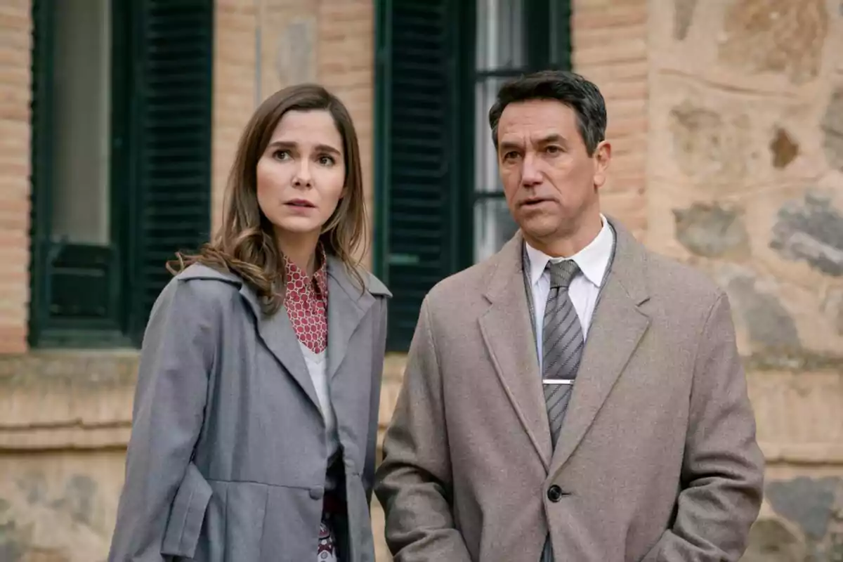 Dues persones amb expressió seriosa i abrics d’hivern, que són l’Oriol Tarrasón i la Natalia Sánchez, estan dretes davant d’un edifici de maó i pedra amb finestres fosques. Dues persones amb expressió seriosa i abrics d’hivern, que són l’Oriol Tarrasón i la Natalia Sánchez, estan dretes davant d’un edifici de maó i pedra amb finestres fosques.