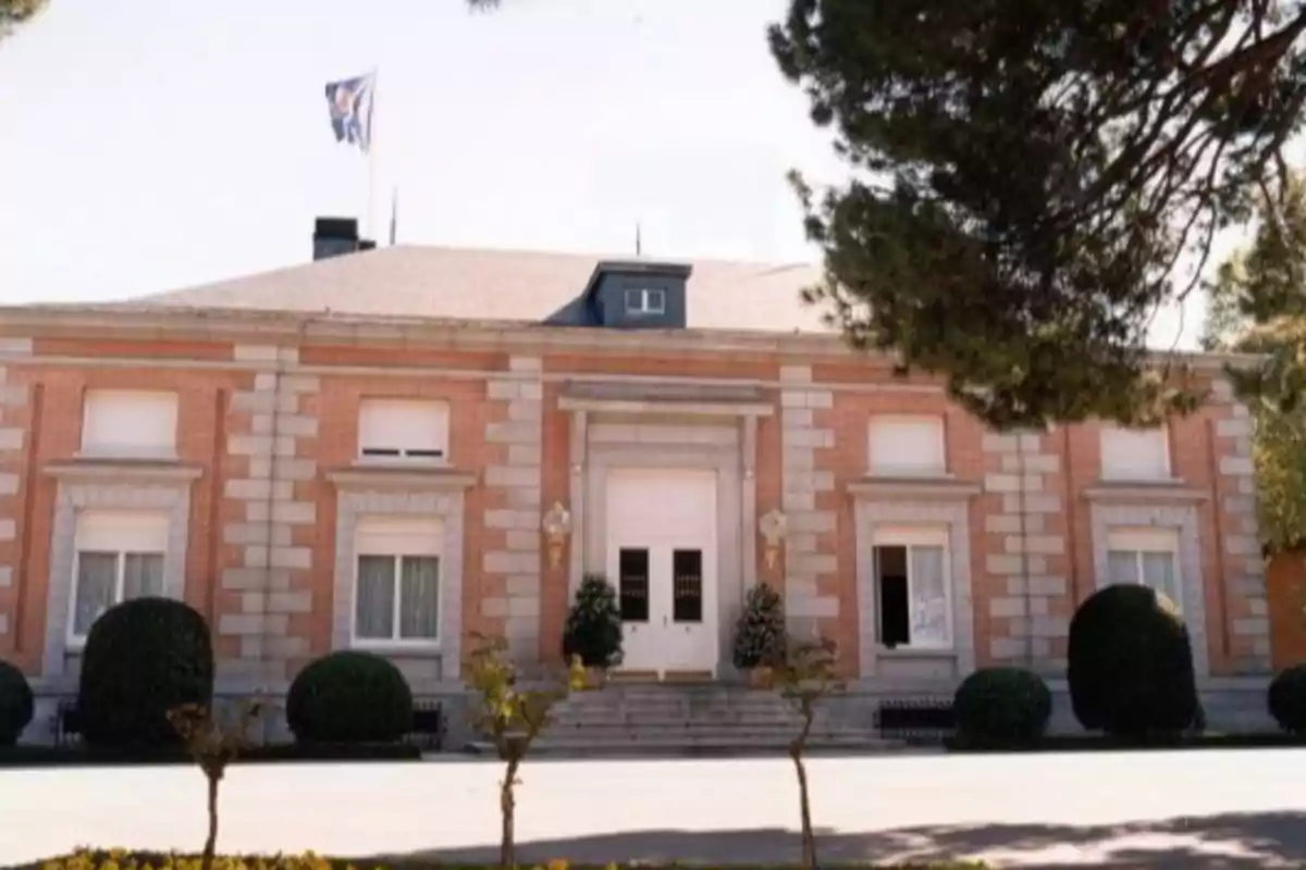 Facade of a brick building with white windows and a flag waving on the roof, surrounded by trees and trimmed bushes Facade of a brick building with white windows and a flag waving on the roof, surrounded by trees and trimmed bushes