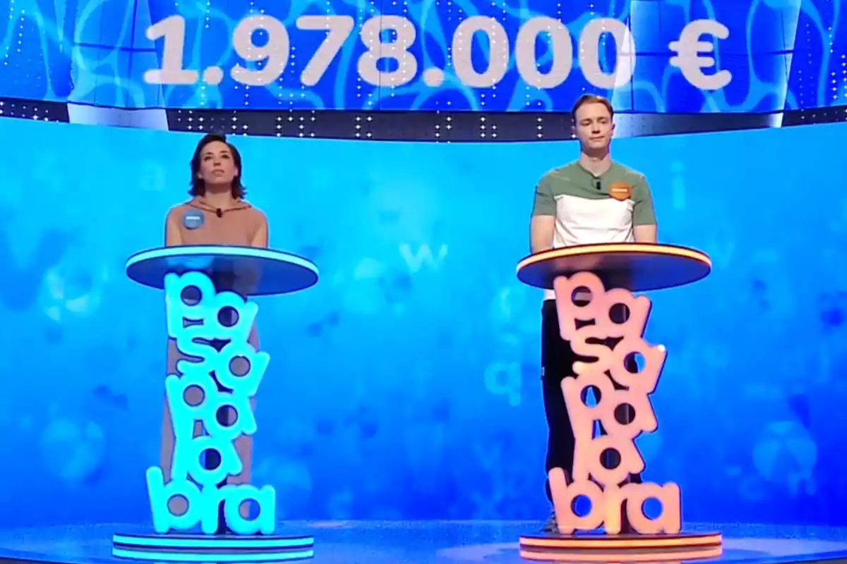 Rosa i Manu drets en un plató de televisió amb decoració blava i taronja, cadascun darrere d’un faristol amb la paraula Rosa i Manu drets en un plató de televisió amb decoració blava i taronja, cadascun darrere d’un faristol amb la paraula