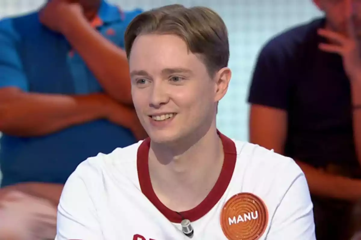 Manu somrient amb samarreta blanca i distintiu que diu Manu participant en un programa de televisió amb persones assegudes al fons. Captura de 'Pasapalabra'