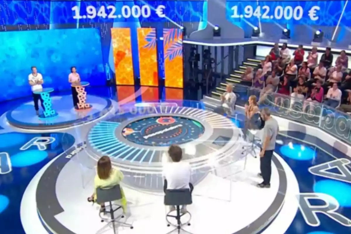Un plató de concurs televisiu amb dos concursants drets davant de faristols, un panell circular al centre, públic assegut al fons i una pantalla que mostra una quantitat de diners en euros. Captura de 'Pasapalabra' Un plató de concurs televisiu amb dos concursants drets davant de faristols, un panell circular al centre, públic assegut al fons i una pantalla que mostra una quantitat de diners en euros. Captura de 'Pasapalabra'