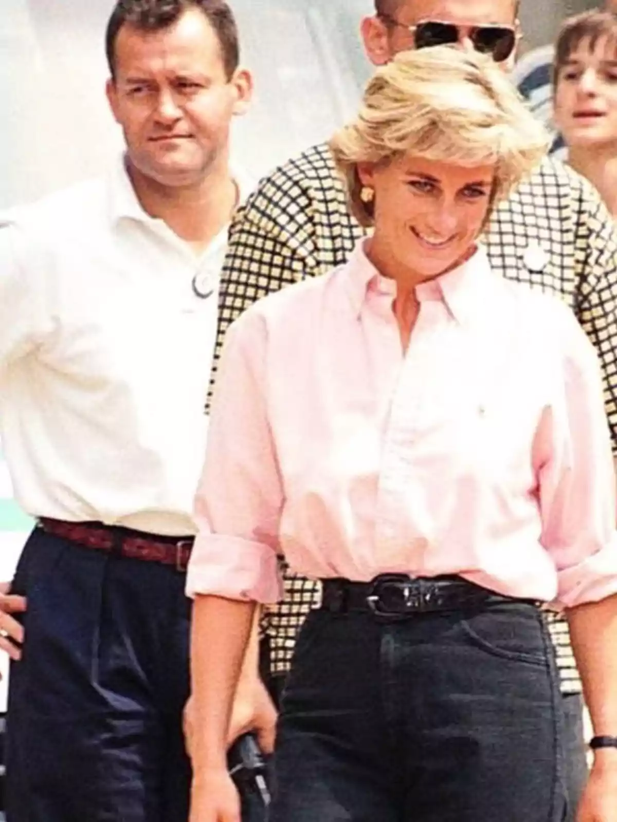 Blonde woman wearing a pink jersey and dark pants smiles while walking accompanied by several people in an outdoor setting.
