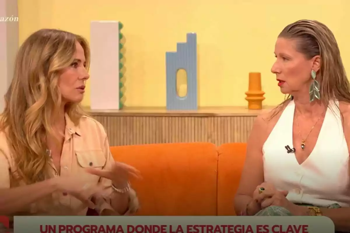 Paula Vázquez y Anne Igartiburu conversan sentadas en un sofá naranja en un set de D Corazón con decoración moderna y fondo amarillo