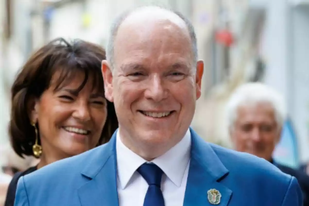 El príncipe Alberto de Mónaco vestido con traje azul sonriendo mientras una mujer sonriente está detrás de él en un entorno urbano desenfocado. El príncipe Alberto de Mónaco vestido con traje azul sonriendo mientras una mujer sonriente está detrás de él en un entorno urbano desenfocado.